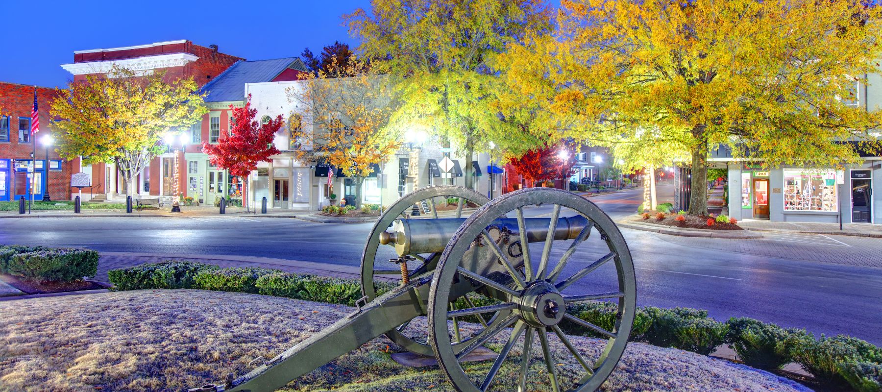 A historic town square at twilight, illuminated by street lights. Autumn leaves in vibrant yellows and reds contrast with the quaint storefronts. In the foreground, a cannon on display signifies historical significance, adding a sense of heritage to this serene setting