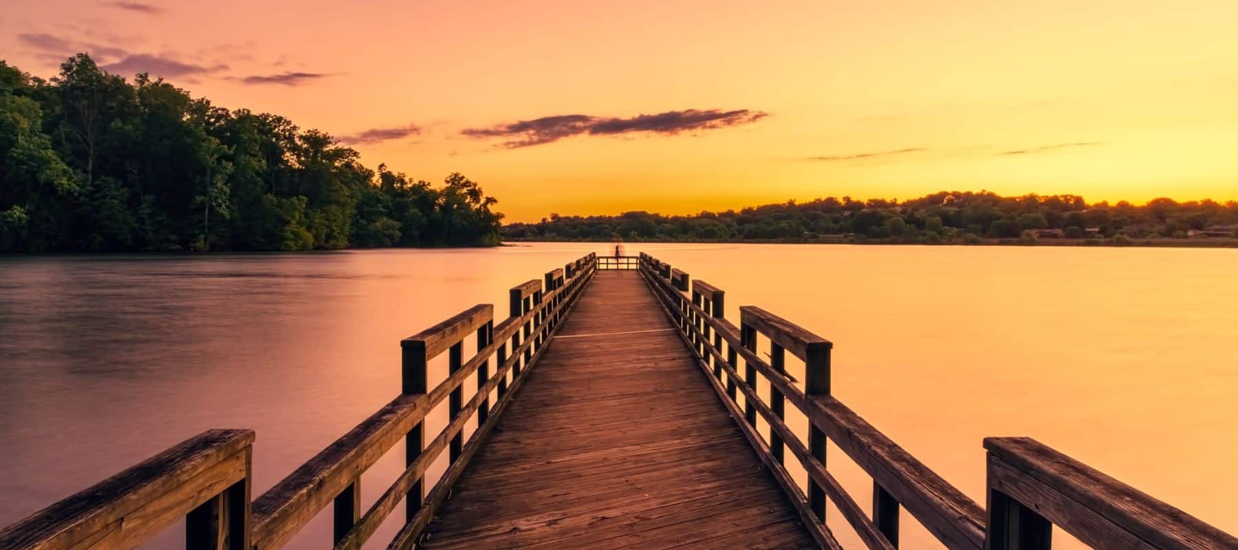 A wooden boardwalk extends out into a calm lake, with the sun setting in the background, casting a warm golden glow over the water and sky. The boardwalk is flanked by railings on both sides, leading to a viewpoint surrounded by lush trees on either side of the lake. The tranquil scene captures the peaceful ambiance of a sunset over the serene water, with hues of orange, yellow, and pink blending in the sky