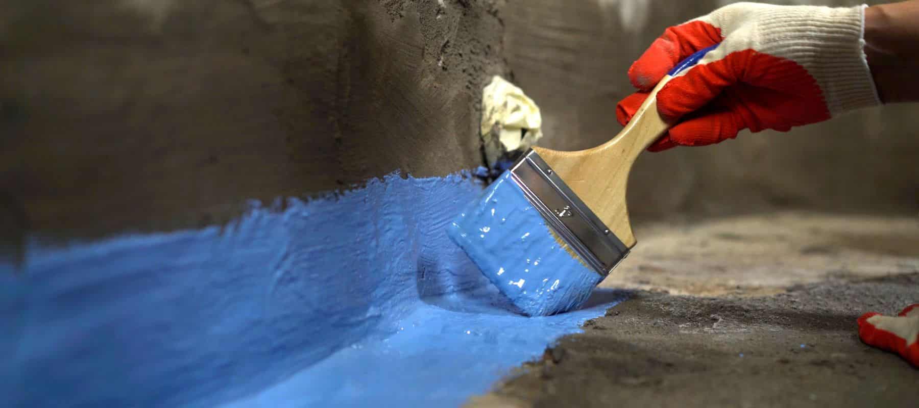 Close-up of a hand applying waterproofing sealant to a foundation wall with a brush