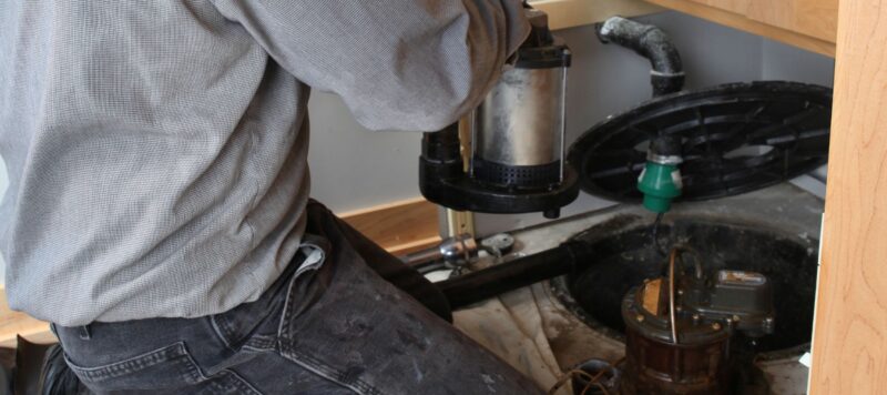 A person kneels beside a sump pump system under a cabinet, preparing for emergency repair