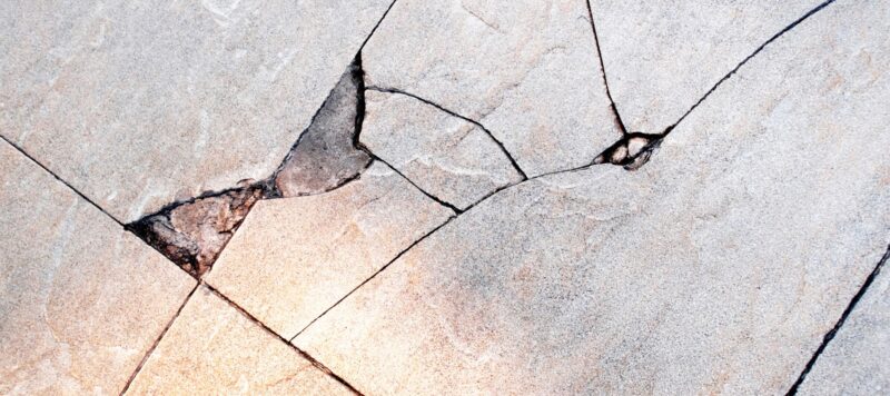 A close-up of a floor shows multiple light-colored tiles with severe, jagged cracks and missing pieces